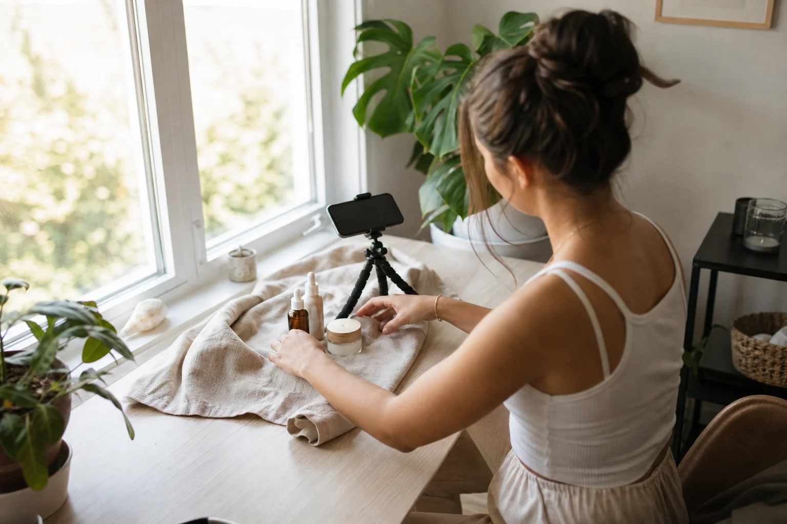 A creator sitting near a bright window with a phone propped on a small tripod, framing a natural-light flat-lay photo of skincare products on a desk