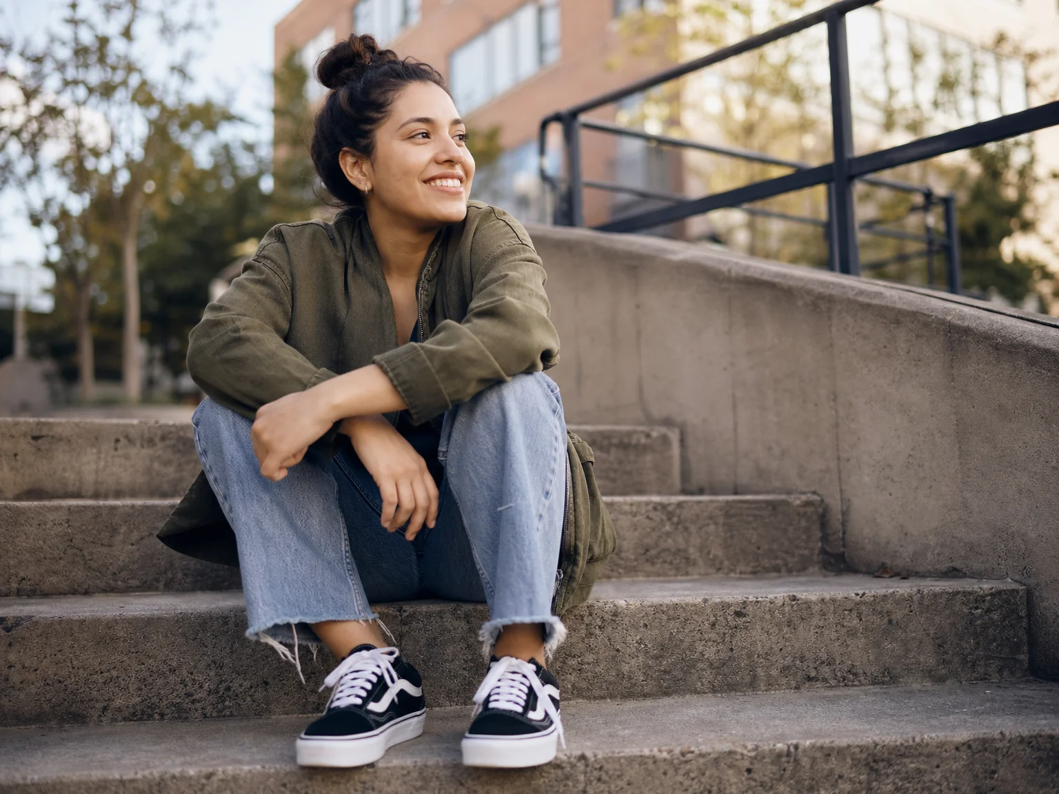 Woman sitting on outdoor concrete steps, ankles crossed, leaning forward with forearms on her knees, photographed from a low angle at her seated eye level