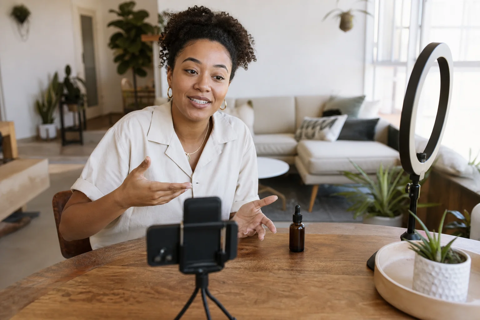 A creator filming themselves with a phone on a tabletop tripod in a bright, naturally lit living room, a product on the table beside them