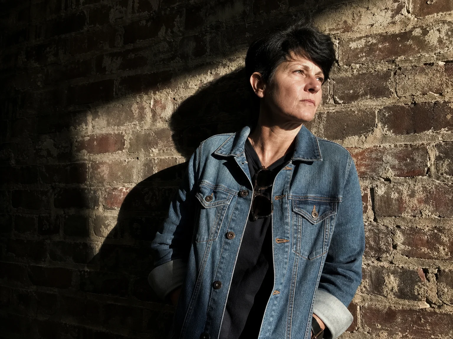 An adult leans against a heavily textured brick wall in strong side light, deep shadows contrasting with the texture of the brick and a wrinkled denim jacket