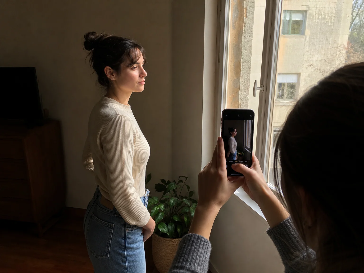 A friend holds up a smartphone to photograph someone standing at a 45-degree angle to a large window, soft daylight lighting one side of their face while the other falls into shadow