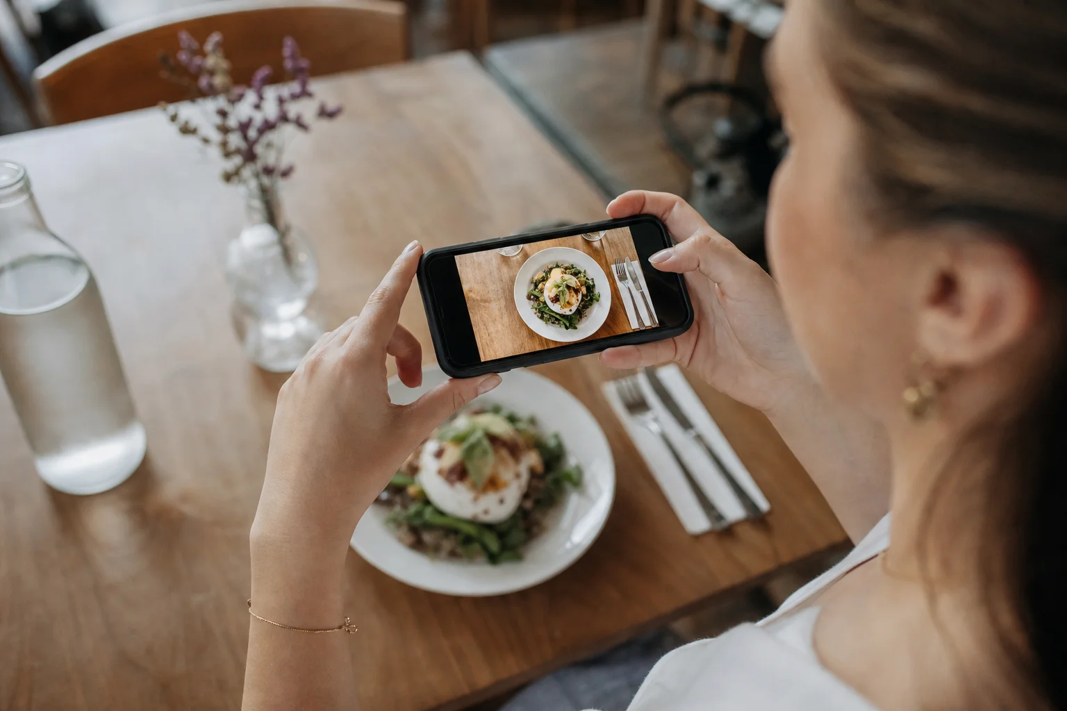 A content creator holding a phone directly above a plate of food at a restaurant table, framing an overhead flat-lay shot