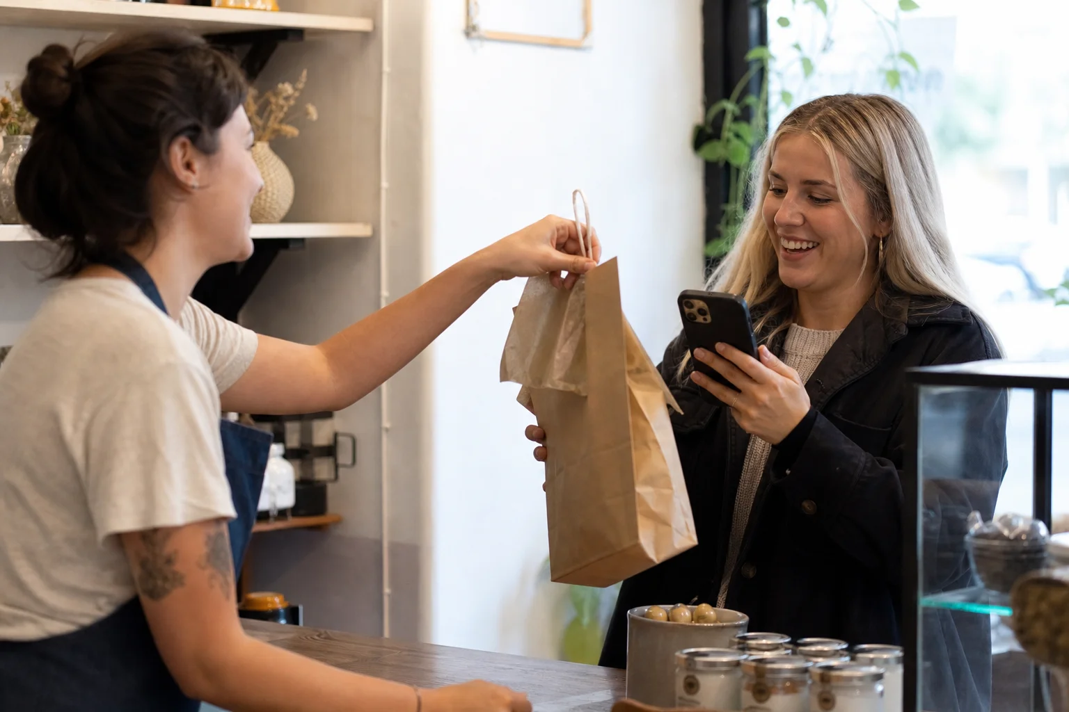 A small business owner in an apron handing a kraft-paper gift bag of products to a content creator holding a phone, mid-unboxing at a shop counter