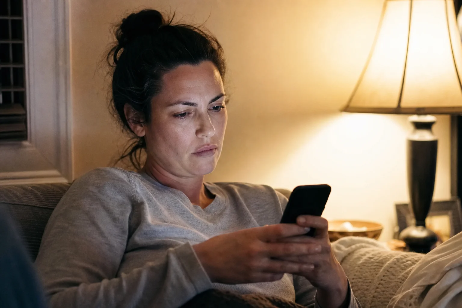 An adult sits alone on a couch at home in the evening, looking at their phone screen with a wary expression