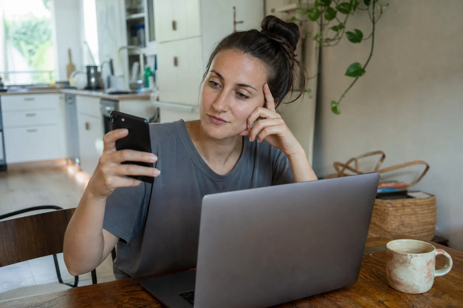 A woman at her kitchen table in natural daylight looking at her phone next to an open laptop, comparing a photo across both screens