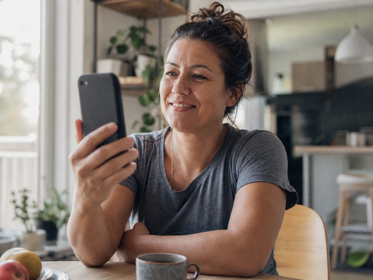 An adult sits at a kitchen table smiling while holding up a smartphone as if on a video call
