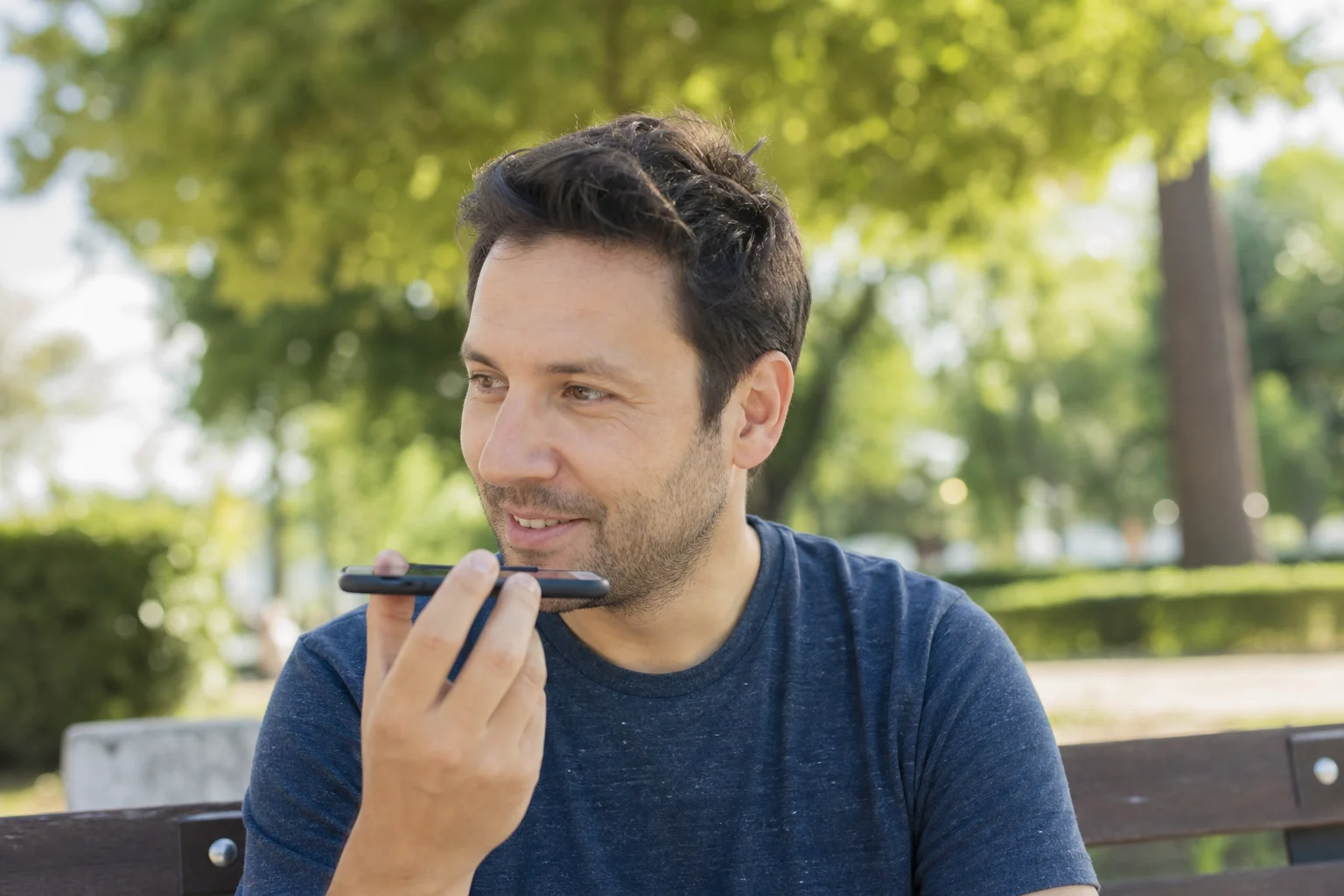 A man on a park bench in late-afternoon light smiling while recording a voice message on his phone