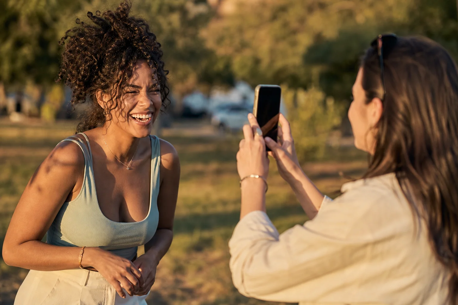 Two friends outdoors at golden hour, one holding up a smartphone taking a candid photo of the other mid-laugh