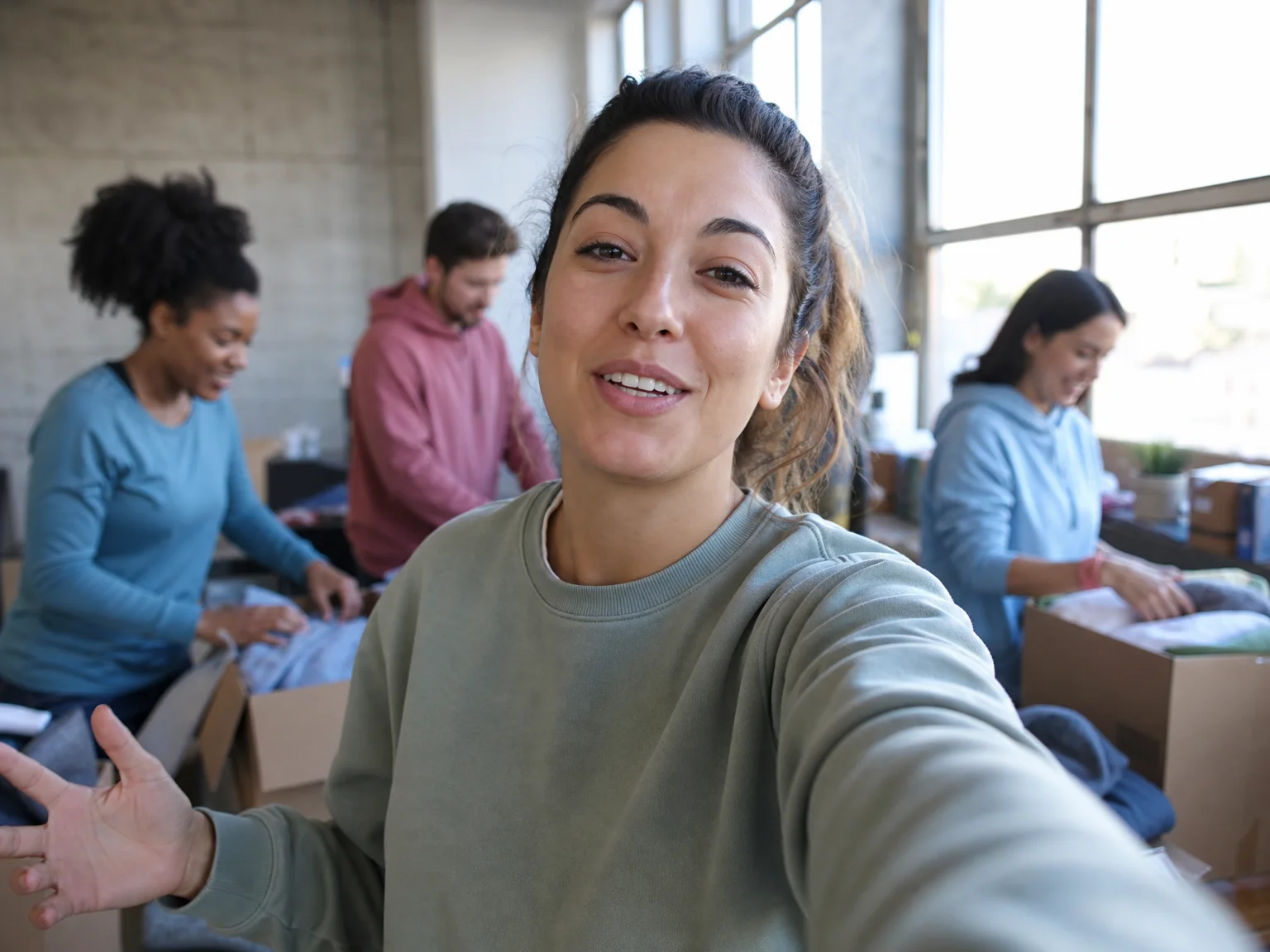 A content creator filming a selfie video at a community volunteer event, with blurred volunteers sorting donation boxes in the background