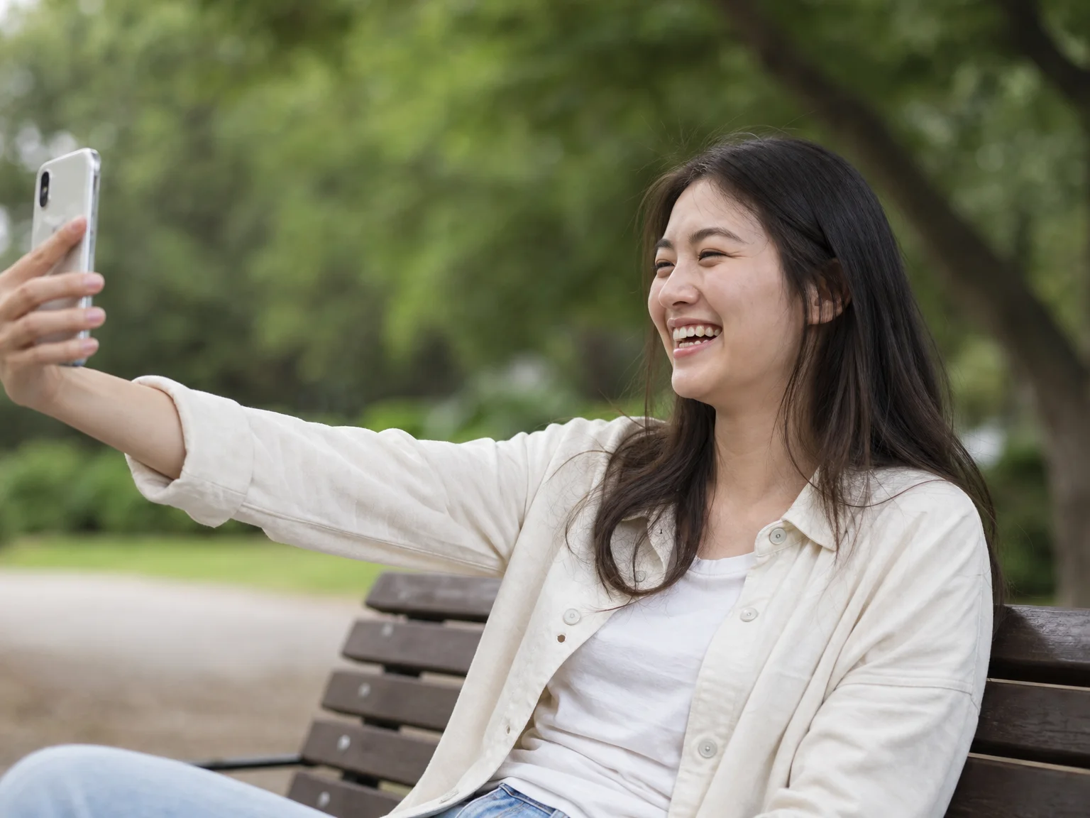 Woman on a park bench pressing her phone's volume button to trigger the shutter during a selfie, mid-laugh